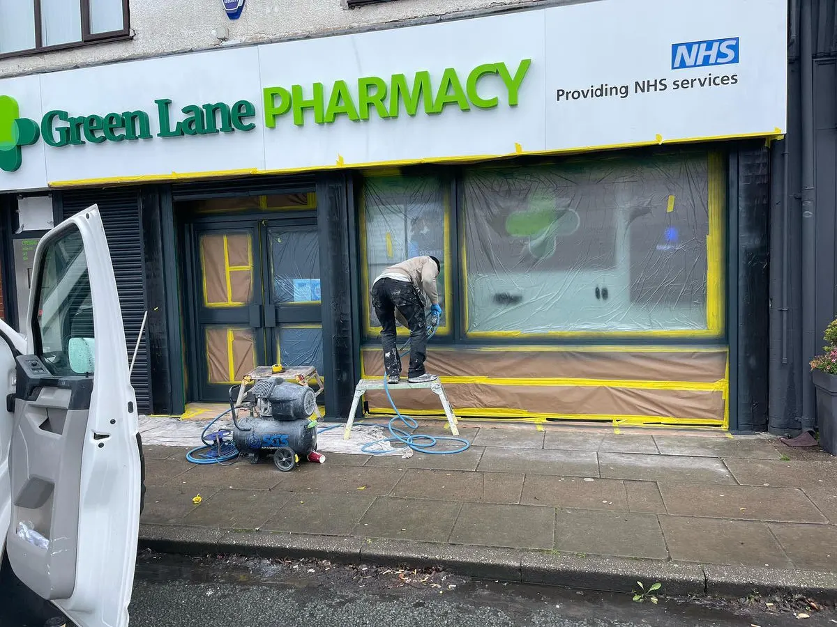 Painter spray-painting a pharmacy shopfront exterior in dark grey with windows masked off