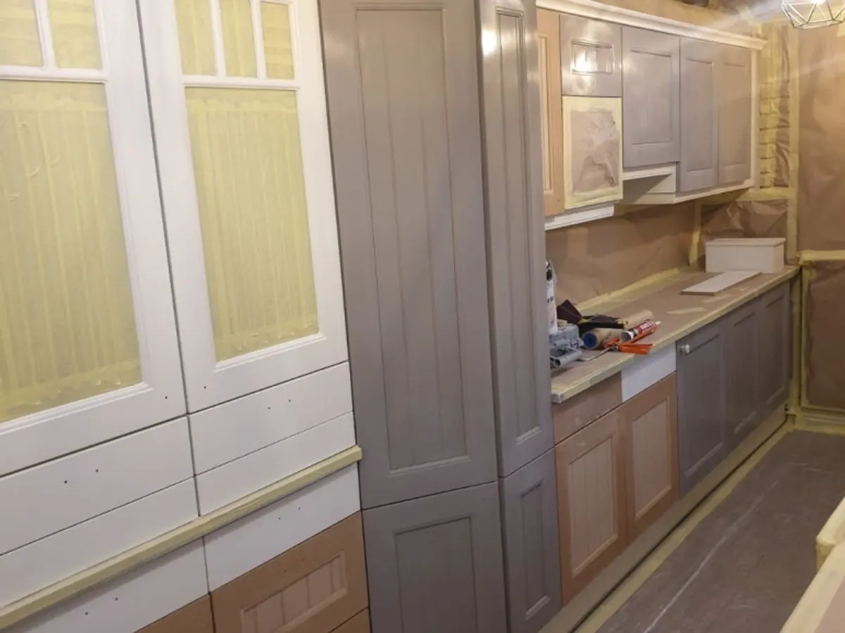 Kitchen cabinets mid-spray with grey primer coat applied and walls masked with protective sheeting