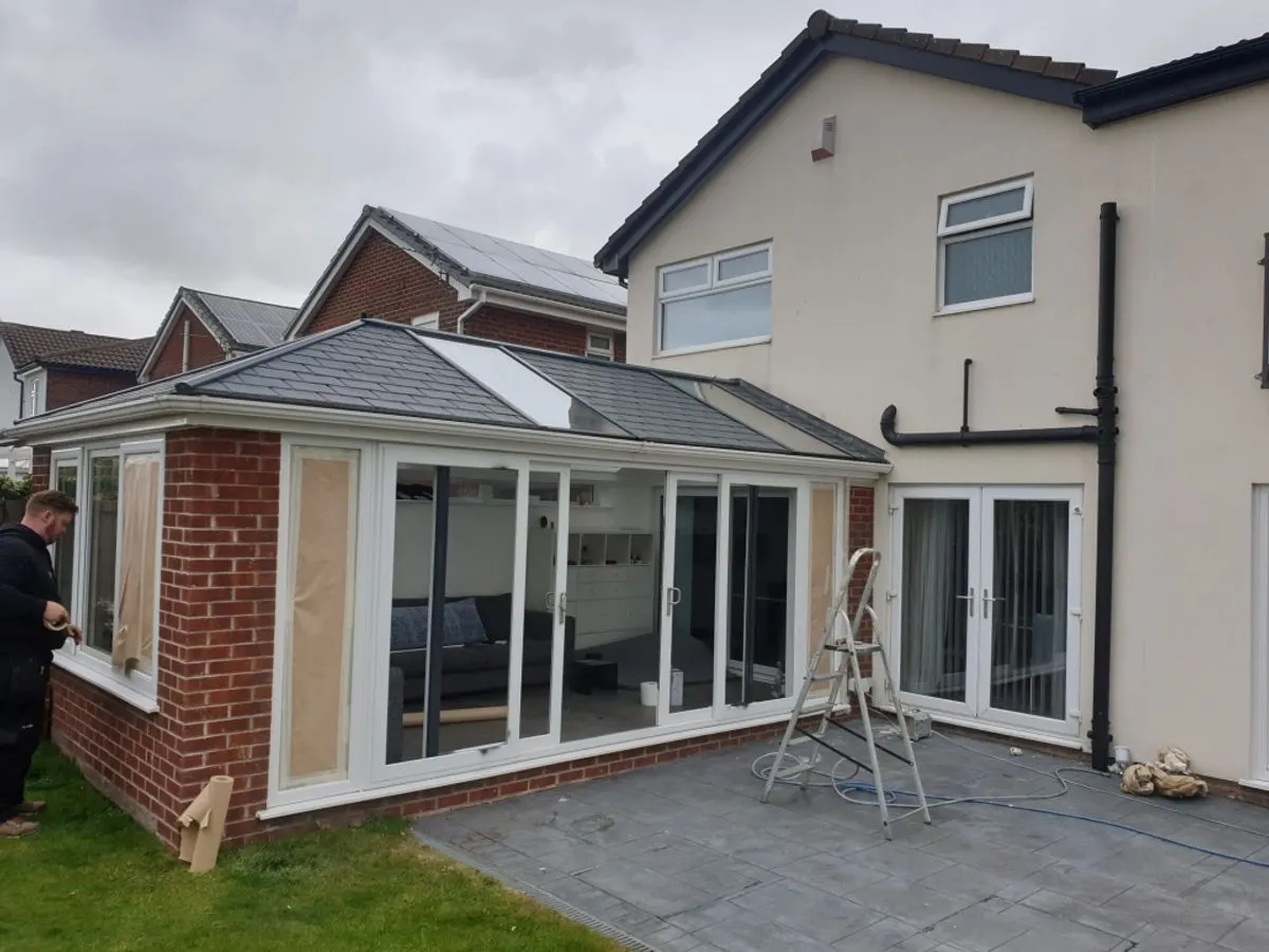 Rear of house with white UPVC conservatory windows being spray-painted by decorator on patio